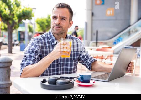 Reifer Freiberufler, der mit einem Laptop sitzt und Orangensaft im Café im Freien trinkt Stockfoto