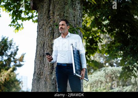 Geschäftsmann hält Smartphone und Solaranlage vor dem Baum Stockfoto