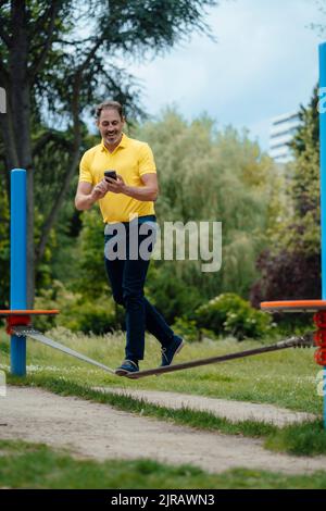 Glücklicher Mann mit Smartphone balanciert auf Drahtseilakt im Park Stockfoto