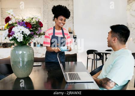 Lächelnde Kellnerin serviert dem Gast Kaffee, der mit einem Laptop im Café sitzt Stockfoto