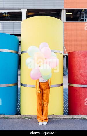 Junge Frau bedeckt das Gesicht mit bunten Luftballons vor den Rohren Stockfoto