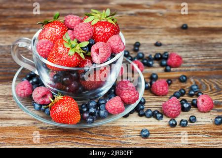 Mix aus frischen reifen Beeren in einer transparenten Tasse auf braunem Holzhintergrund mit Platz für Text. Erdbeere, Heidelbeere im Sortiment. Stockfoto