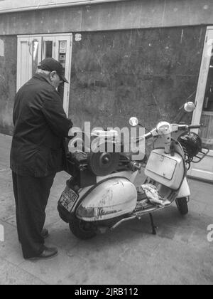 Eine vertikale Graustufenaufnahme eines Mannes, der hinter dem italienischen Vespa-Roller steht Stockfoto