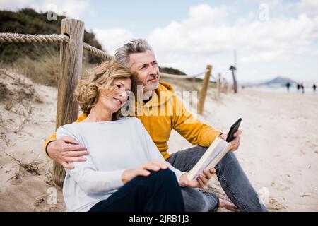 Lächelnder reifer Mann mit Frau, die am Strand ein Buch liest Stockfoto