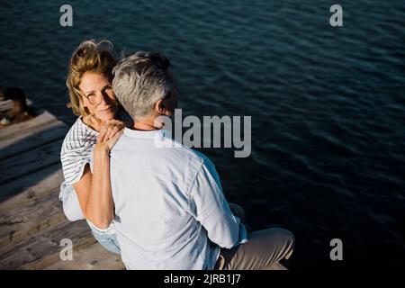 Lächelnde reife Frau, die am Steg mit der Hand auf der Schulter des Mannes sitzt Stockfoto