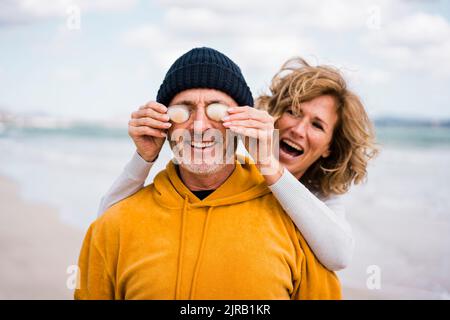 Fröhliche Frau, die die Augen des Mannes mit Muscheln am Strand bedeckt Stockfoto