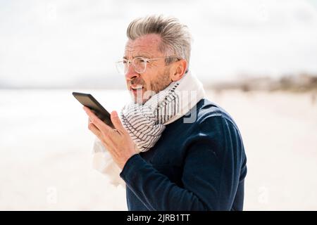 Reifer Mann trägt Schal und redet am Strand mit dem Handy Stockfoto