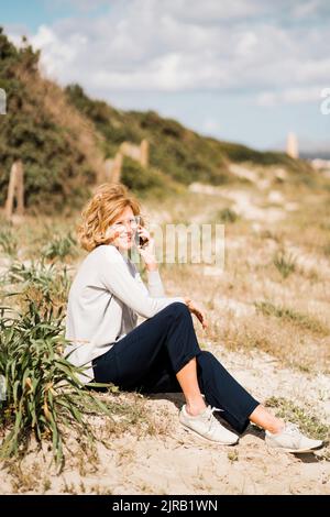 Glückliche, reife Frau, die am Strand am Strand sitzt und mit dem Smartphone spricht Stockfoto