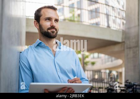 Lächelnder junger Geschäftsmann mit Tablet-PC an der Wand Stockfoto