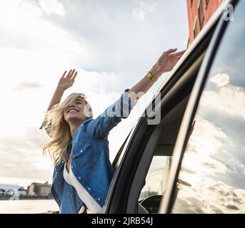 Glückliche junge Frau mit erhobenen Armen, die sich aus dem Autofenster lehnen Stockfoto