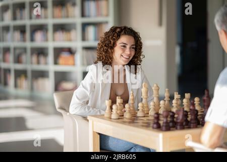 Lächelnde Geschäftsfrau mit lockigen Haaren, die mit einem Kollegen im Büro Schach spielt Stockfoto