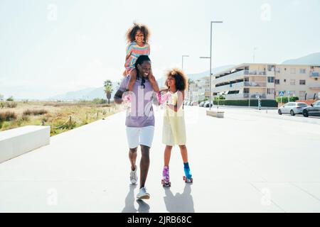 Lächelndes Mädchen Rollschuh vom Vater, der Schwester auf den Schultern trägt Stockfoto