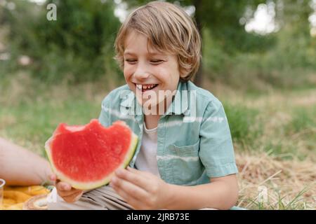 Lächelnder Junge mit Wassermelonenscheibe, der im Park sitzt Stockfoto