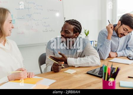 Lächelnde Geschäftsfrau im Gespräch mit einem Geschäftsmann, der von einem Kollegen im Büro sitzt Stockfoto