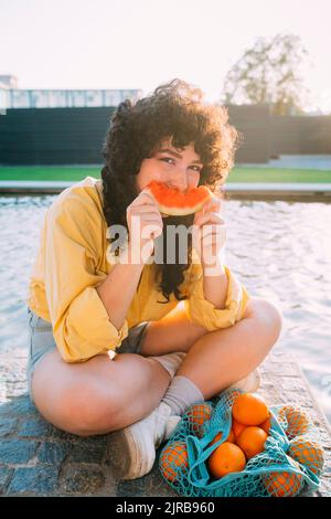 Frau mit Orangen im Netzbeutel, die Wassermelone am Teich isst Stockfoto