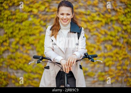 Porträt einer lächelnden modernen Frau in beigem Trenchcoat mit Fahrrad an der grünen Wand im Freien auf der Straße der Stadt. Stockfoto