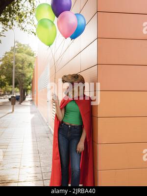 Junge Frau trägt Krokodilmaske und steht mit bunten Ballons an der Wand Stockfoto