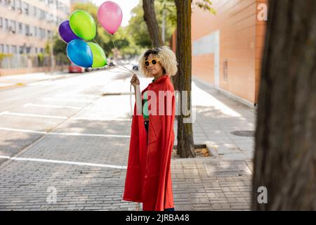 Junge Frau mit Umhang und Sonnenbrille, die mit bunten Luftballons auf dem Fußweg steht Stockfoto