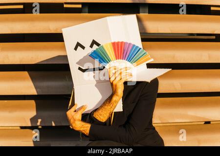 Geschäftsmann trägt Box mit Smiley Gesicht hält Regenbogenfarben Handfächer Stockfoto