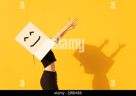 Glückliche Frau trägt Box mit Smiley Gesicht tanzen an der gelben Wand Stockfoto