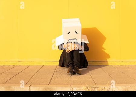 Mann in einer Schachtel mit traurigem Gesicht, der vor einer gelben Wand auf einem Fußweg sitzt Stockfoto