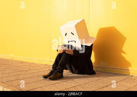 Mann in einer Schachtel mit traurigem Gesicht, der auf dem Fußweg sitzt Stockfoto