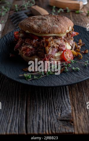 Schweineburger mit gerösteten Zwiebeln, Käse, Gurken und Tomaten auf einem dunklen Teller gezogen Stockfoto