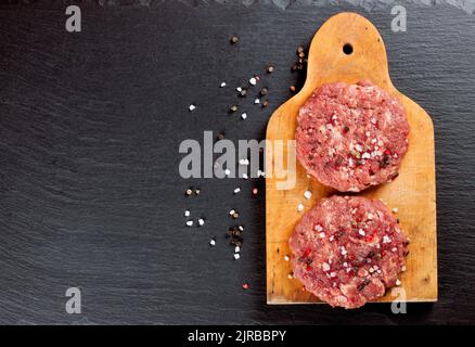 Frischer roher, hausgemachter Hackfleisch-Steak-Burger mit Gewürzen, auf einem Schneidebrett auf einem schwarzen Schiefertisch, Kopierfläche, Draufsicht Stockfoto