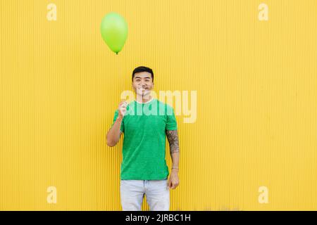 Lächelnder junger Mann, der einen grünen Ballon vor der Wand hält Stockfoto