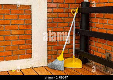 Gelber Besen und Kehrschaufel, die sich an der roten Ziegelwand an der Ecke einer Terrasse des Hauses lehnen. Stockfoto