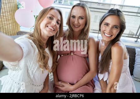 Glückliche Freunde machen Selfie mit einer Schwangeren, die auf dem Sofa sitzt Stockfoto