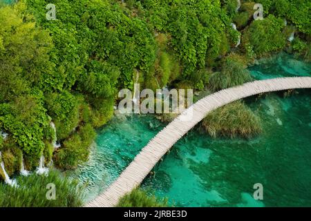 Wandertouristen wandern auf einer hölzernen Fußgängerbrücke, die den See mit türkisfarbenem Wasser im Naturschutzgebiet umgibt. Majestätischer Blick auf den blauen See inmitten malerischer Landschaft auf den Plitvicer Seen Stockfoto