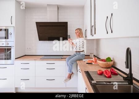 Frau, die zu Hause in der heimischen Küche ein Mobiltelefon benutzt Stockfoto