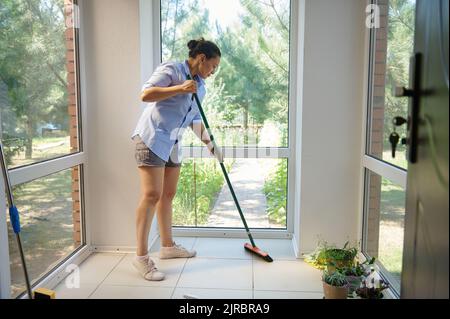 Multiethnische Frau, die Besen benutzt, fegt während einer Hausreinigung den Boden auf der Veranda. Reinigungsservice. Stockfoto