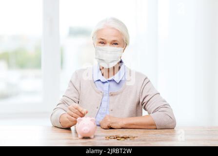 Alte Frau in Maske, die Münze ins Sparschwein legt Stockfoto