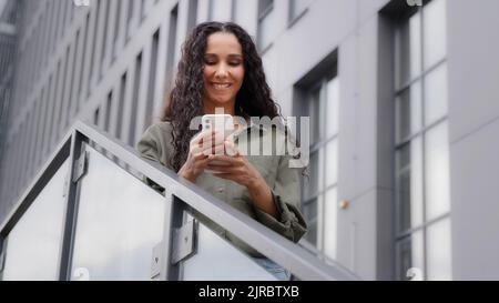 Lächelnd kaukasisch hispanisch glücklich zufrieden Frau Benutzer Dame stehen un Stadt im Freien auf Treppen chatten SMS suchen im Netz mit Handy App Telefon Stockfoto