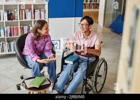 Hochwinkelporträt eines jungen asiatischen Mannes im Rollstuhl, der mit einem Freund spricht, während er in der Bibliothek zusammen studiert Stockfoto