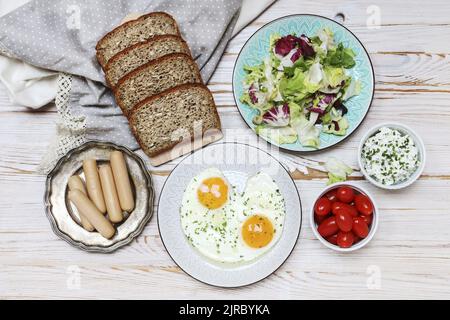Teller mit Spiegeleiern, Brot, Salat, Wurst und Quark auf braunem Holztisch, Kopierplatz. Stockfoto