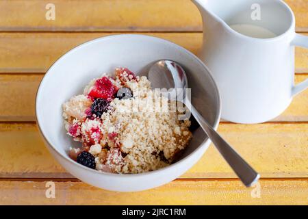 Eine Portion hausgemachter Brombeere und Apfelbrösel in einer Schüssel serviert. Zur traditionellen englischen Fruchtwüste gibt es frische Doppelcreme Stockfoto