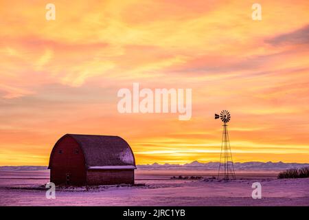 Eine hölzerne Scheune und Windmühle, die von einem Winteruntergang im ländlichen Süden Albertas umragt werden Stockfoto