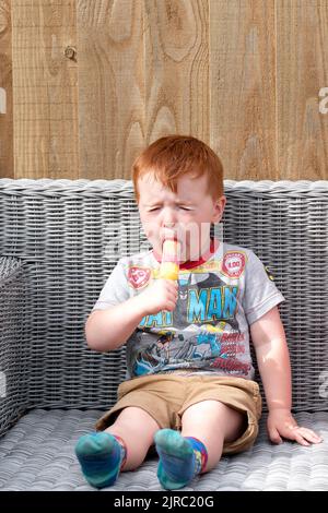 Ein kleines Kind wird gezeigt, wie es einen sauren Eislolly leckt. Der Junge sitzt draußen und grimazt den bitteren Geschmack des aromatisierten Eises. Stockfoto