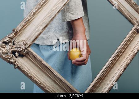 Ein junger Mann aus dem Jahrtausend hält einen Apfel auf der Handfläche in einem vergoldeten Bilderrahmen Stockfoto