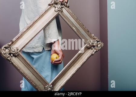 Ein junger Mann aus dem Jahrtausend hält einen Apfel auf der Handfläche in einem vergoldeten Bilderrahmen Stockfoto