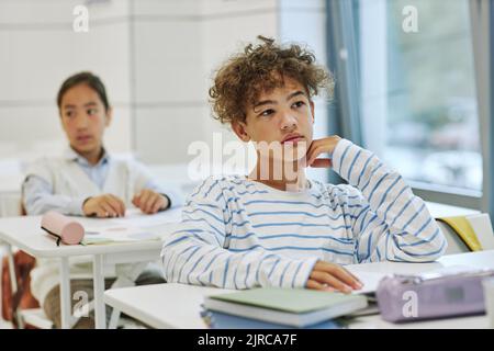 Porträt eines jungen Jungen mit lockigen Haaren, der im Klassenzimmer in minimalem Weiß am Schreibtisch sitzt und Platz zum Kopieren hat Stockfoto