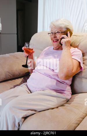 Foto von gealterten Frau glücklich positives Lächeln Gespräch Handy trinken Alkohol Wein Rest entspannen Wochenende zu Hause. Ältere Frau auf dem Sofa telefoniert mit gl Stockfoto