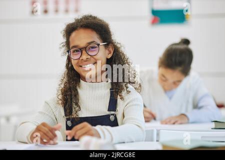 Porträt eines lächelnden schwarzen Schulmädchen mit Brille und Blick auf die Kamera, während sie am Schreibtisch in der Schule sitzt, Platz kopieren Stockfoto