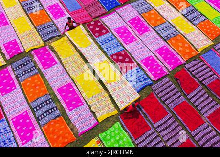 Narayanganj, Bangladesch. 23. August 2022. Farbenfrohe Stoffstreifen bilden ein auffälliges Display, da sie in ordentlichen Reihen auf einem Feld in Narayanganj, Bangladesch, angeordnet sind. Die langen Baumwolltücher werden lokal als ''Saree'' bezeichnet - ein traditionelles Kleidungsstück für Frauen, das unter der heißen Sonne trocknen soll und mit hellen Farben gefärbt wurde. Hier werden täglich etwa 4000 Stück Stoff zum Trocknen gelegt. Der Prozess dauert in der Regel drei Stunden, wobei jeder Satz von 200 Stück auf einmal bei Temperaturen trocknen kann, die über 36 Grad celsius erreichen können. Schön verzierte bunte lange Tücher entstehen Stockfoto