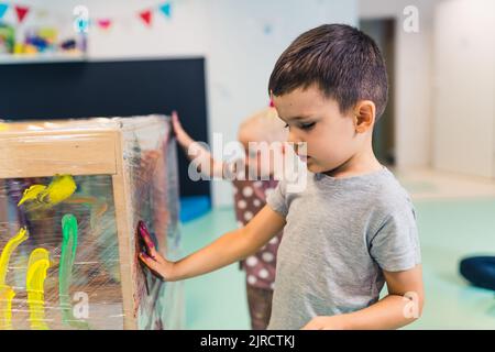 Frischhaltefolie Malerei für die Verbesserung der Kinder Phantasie und Gehirnentwicklung. Kleinkinder malen mit Tempera-Farben auf einem Frischhaltefilm, der um den hölzernen Regalständer gewickelt ist. Lustige Aktivität für Kinder feine und grobe Motorik im Kindergarten. Hochwertige Fotos Stockfoto