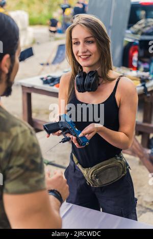 Kaukasische junge Frau mit langen Haaren, die Sicherheitskopfhörer trägt und Handfeuerwaffen vom Mann abgibt. Kauf und Verkauf von Schusswaffen Thema. Vertikale Aufnahme im Freien. Hochwertige Fotos Stockfoto