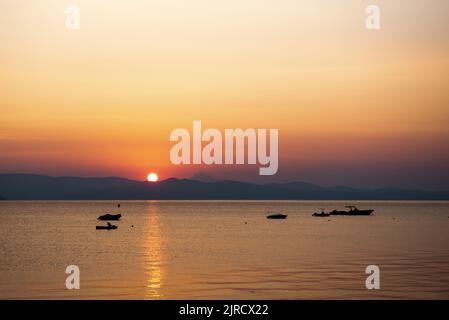 Sonnenaufgang über den Sithonia-Bergen von Kassandra aus gesehen in Halkidiki Griechenland. Die Silhouetten des Boots liegen auf dem ruhigen blauen Meer. Stockfoto
